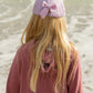 A child with long blonde hair, wearing a mauve sweater and the Bits & Bows Blonde Mermaid Bow Baseball Hat, stands facing the ocean, ready for summer sportswear adventures.