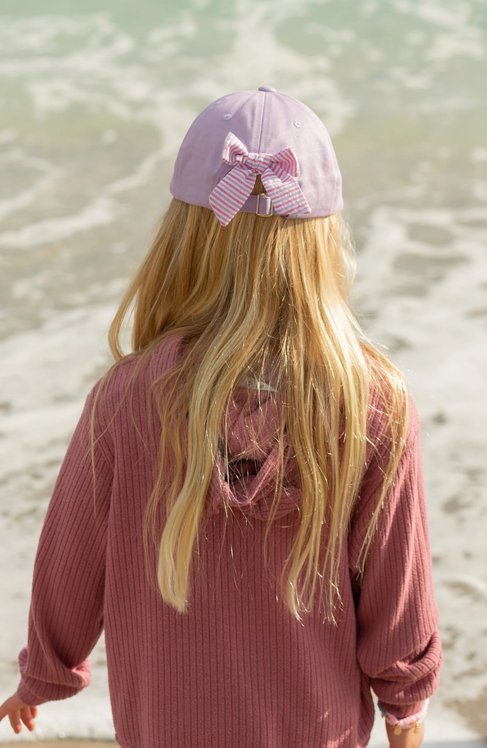 A child with long blonde hair, wearing a mauve sweater and the Bits & Bows Blonde Mermaid Bow Baseball Hat, stands facing the ocean, ready for summer sportswear adventures.