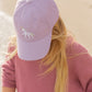 A person with long blonde hair wears the Bits & Bows Rainbow Unicorn Bow Baseball Hat and a matching ribbed sweater, standing near a sandy beach.