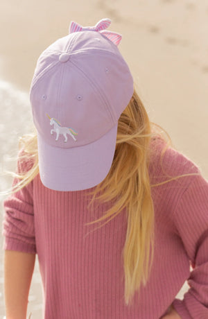 A person with long blonde hair wears the Bits & Bows Rainbow Unicorn Bow Baseball Hat and a matching ribbed sweater, standing near a sandy beach.