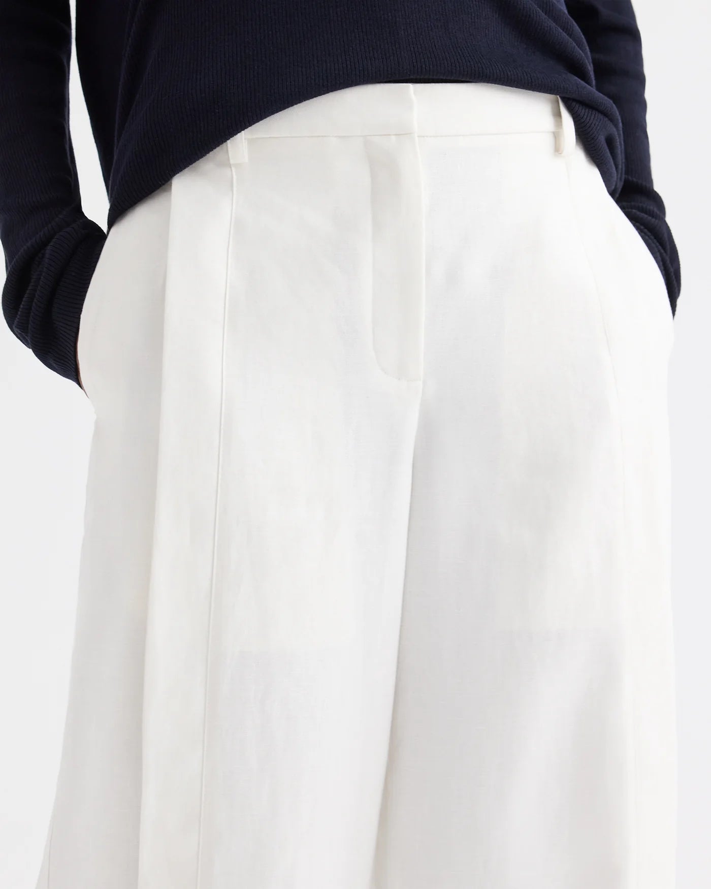 A person wearing a navy long-sleeve top tucked into the TWP Taite Chintzed Linen Pant—a pair of high-rise, wide-leg ivory trousers—with hands in pockets.