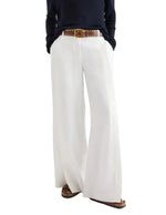 A person shown from the shoulders down wears a navy long-sleeve top, brown belt, and the TWP Taite Chintzed Linen Pant in high-rise ivory, hands in pockets.