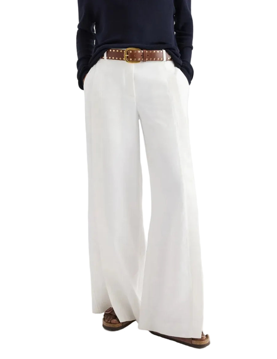 A person shown from the shoulders down wears a navy long-sleeve top, brown belt, and the TWP Taite Chintzed Linen Pant in high-rise ivory, hands in pockets.