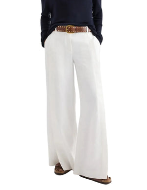 A person shown from the shoulders down wears a navy long-sleeve top, brown belt, and the TWP Taite Chintzed Linen Pant in high-rise ivory, hands in pockets.