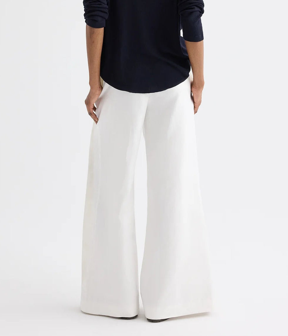 A person stands with their back to the camera, wearing a navy long-sleeve top and TWP’s Taite Chintzed Linen Pant—high-rise, wide-leg ivory pants—against a plain background.