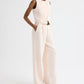 A person wears the Hudson St Pant by TWP—a sleeveless, light beige jumpsuit made from coated viscose linen—featuring wide-leg styling, a relaxed mid-rise silhouette with a belt, and paired with sandals, standing against a plain white background.