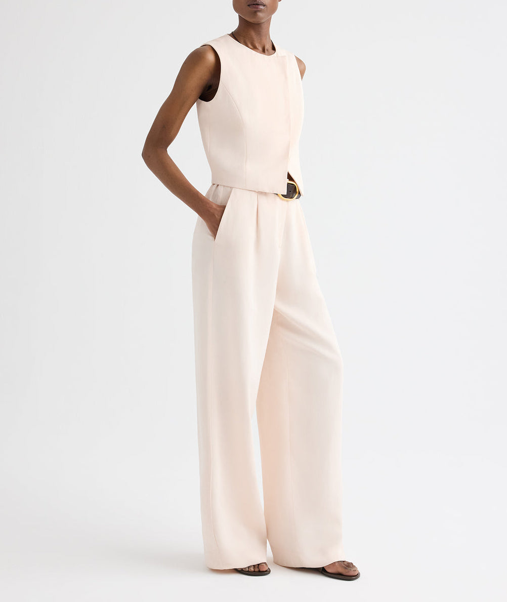 A person wears the Hudson St Pant by TWP—a sleeveless, light beige jumpsuit made from coated viscose linen—featuring wide-leg styling, a relaxed mid-rise silhouette with a belt, and paired with sandals, standing against a plain white background.
