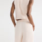 A person stands with hands in pockets, shown from the back against a plain background, wearing a sleeveless beige top paired with TWP's high-waisted Hudson St Pant in coated viscose linen featuring a wide-leg silhouette.