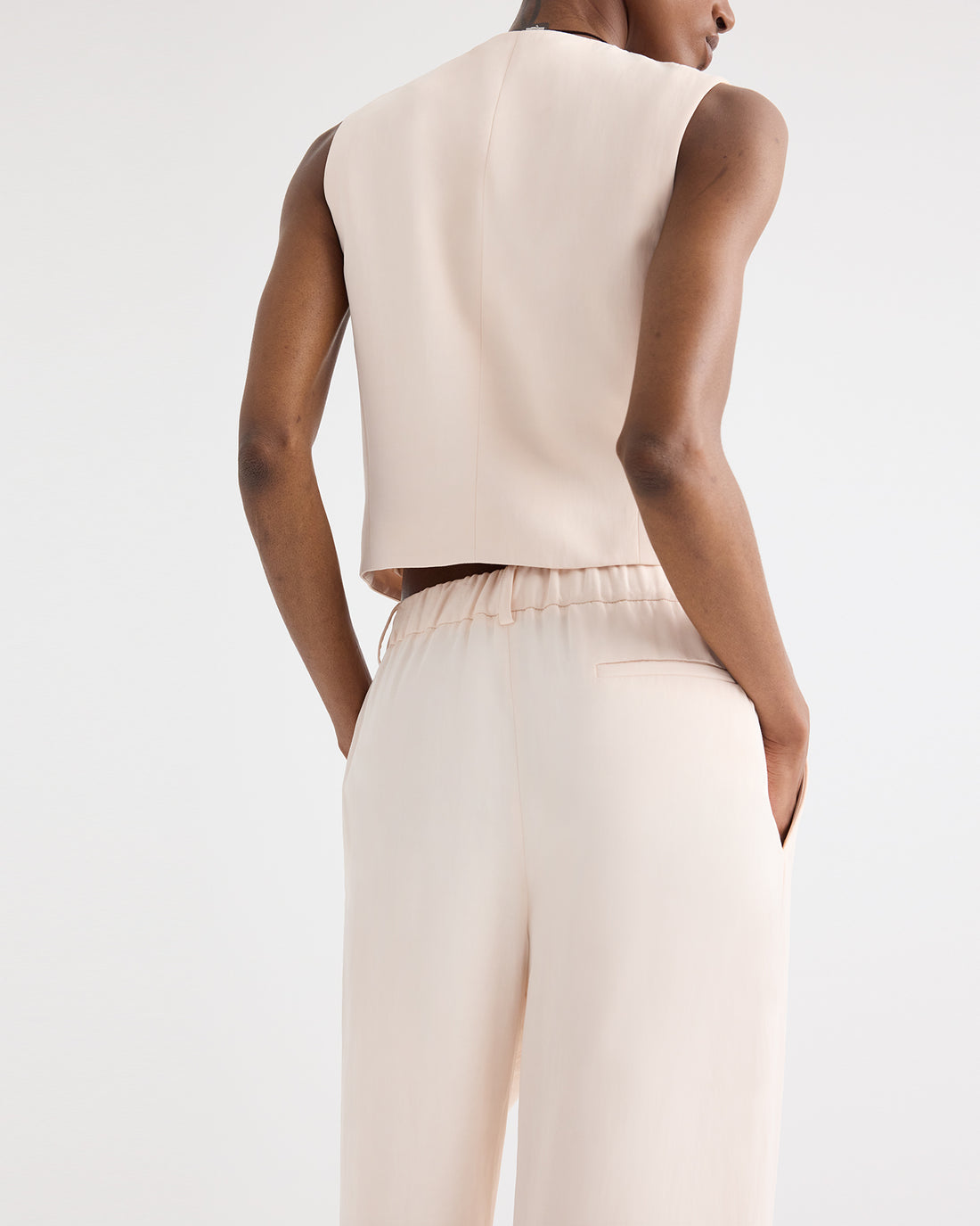 A person stands with hands in pockets, shown from the back against a plain background, wearing a sleeveless beige top paired with TWP's high-waisted Hudson St Pant in coated viscose linen featuring a wide-leg silhouette.
