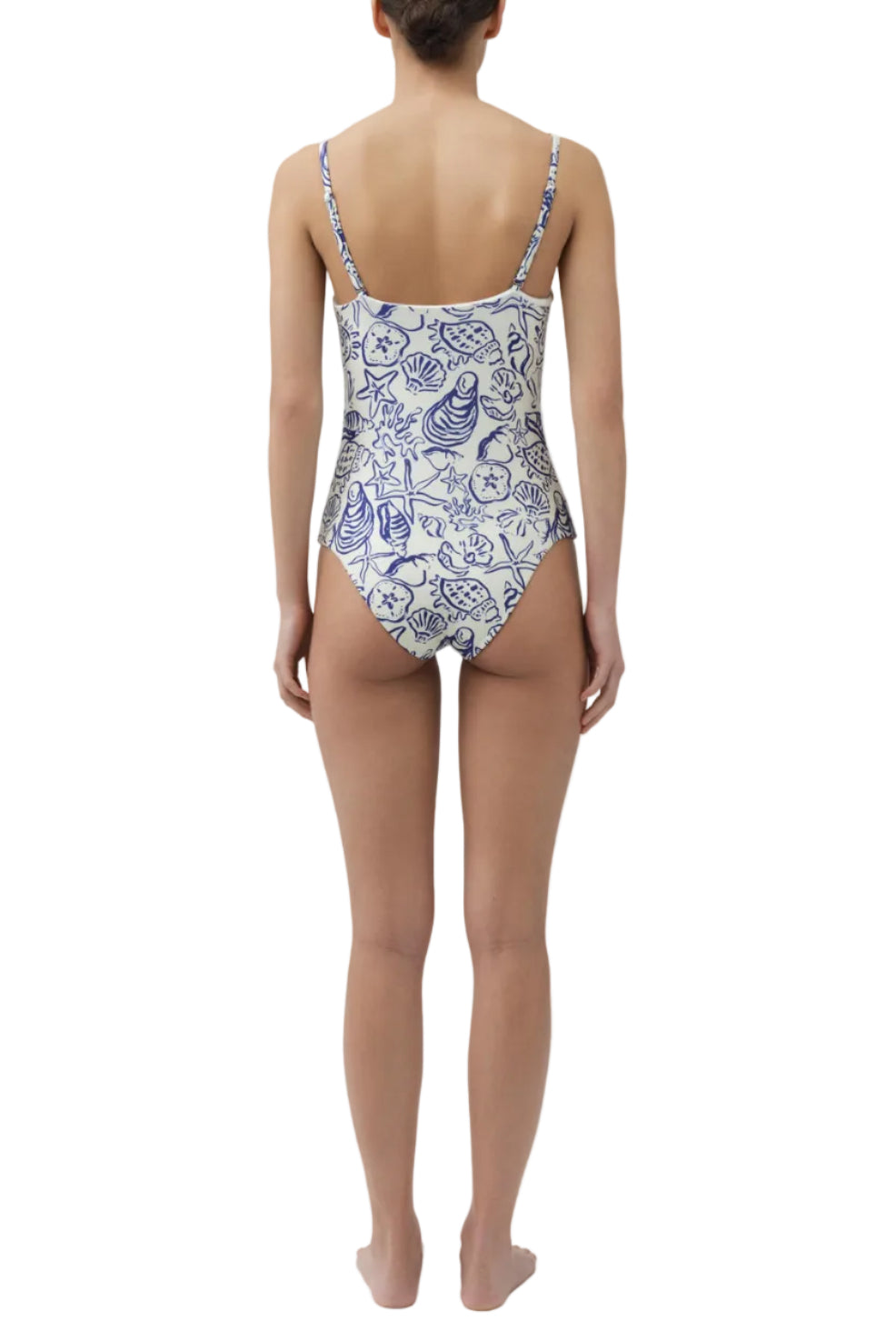 A woman faces away, wearing the Simkhai Ottilie Strapless One Piece—a white swimsuit featuring blue seashell motifs and ocean-themed patterns.