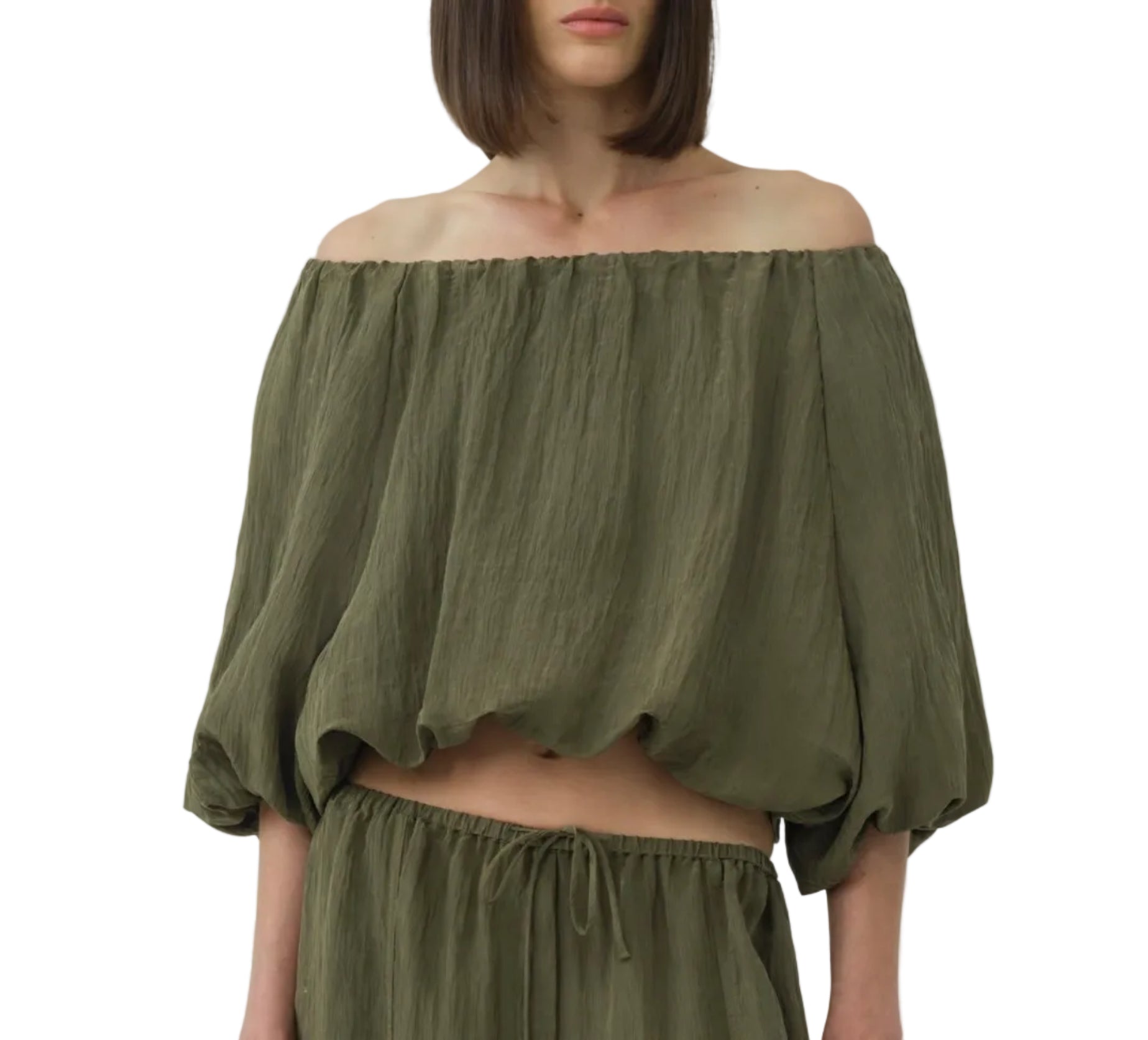 A woman wears the Simkhai Anni Puff Sleeve Cropped Top, an off-the-shoulder, loose-fit olive green blouse in lightweight crinkled fabric, paired with matching drawstring pants against a white background.