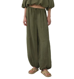 Model wears the Simkhai Freya Parachute Pant, featuring a loose fit, olive green hue, drawstring waist, and elastic cuffs, styled with a coordinating top and beige sandals.