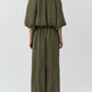 A person stands facing away, wearing the Simkhai Anni Puff Sleeve Cropped Top in olive green with gathered neckline and puff sleeves, styled with beige sandals against a plain light background.