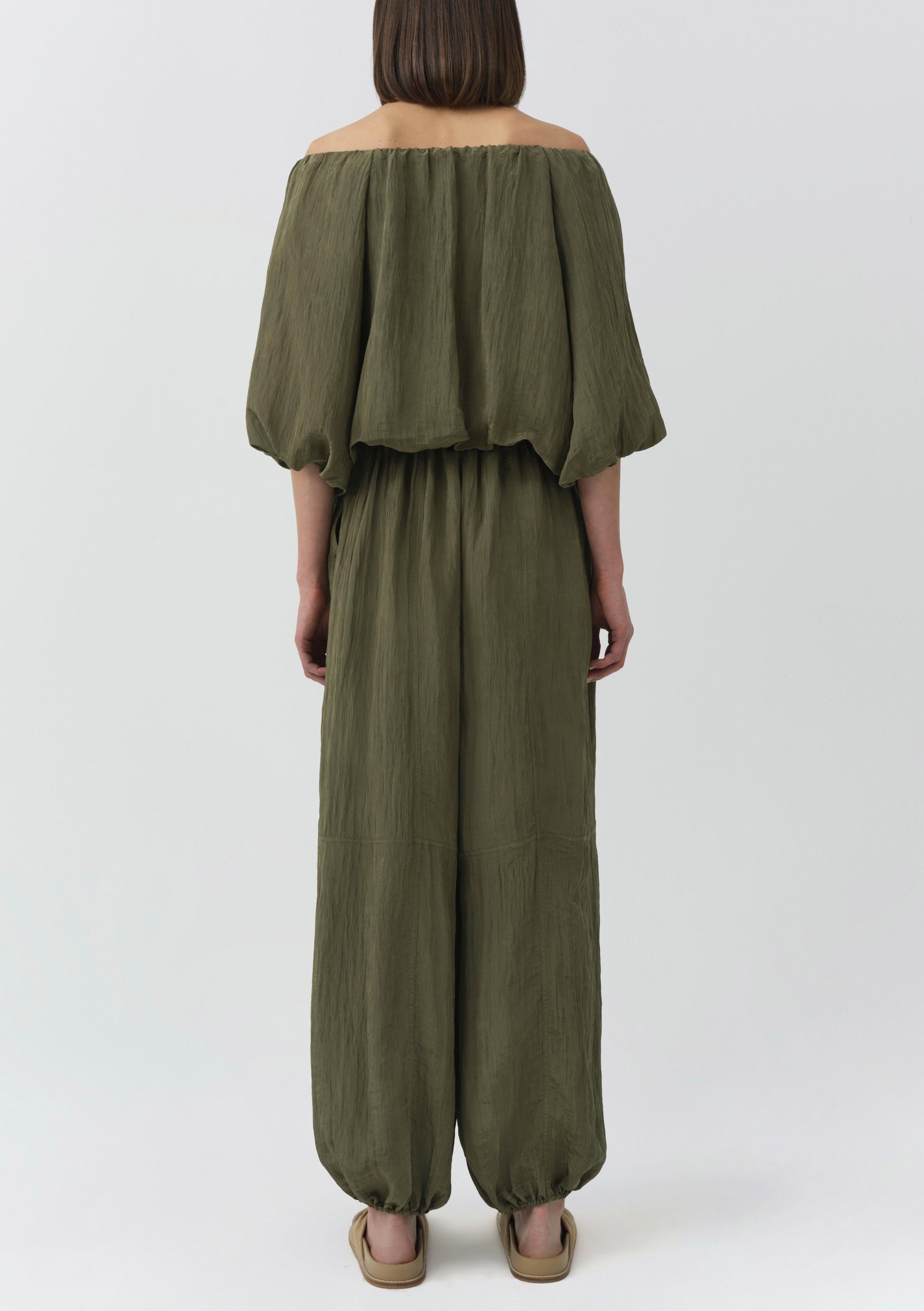 A person stands facing away, wearing the Simkhai Anni Puff Sleeve Cropped Top in olive green with gathered neckline and puff sleeves, styled with beige sandals against a plain light background.