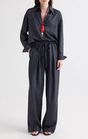 A person stands against a plain white background, dressed in TWP’s PJ Evening Stripe Silk Pant and a matching loose-fitting dark pinstripe shirt, accessorized with a red lanyard and keys.