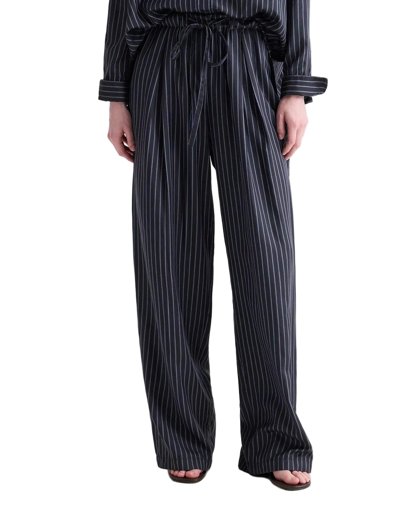 A person wears the TWP PJ Evening Stripe Silk Pant—loose-fitting, dark pinstriped trousers with a drawstring waist reminiscent of pajama-inspired styling—standing with feet slightly apart and hands relaxed at their sides.