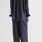A person stands with their back to the camera, dressed in the TWP PJ Evening Stripe Silk Pant set—a loose-fitting, dark ensemble featuring wide-leg pants and a long-sleeved top adorned with a subtle evening stripe pattern.