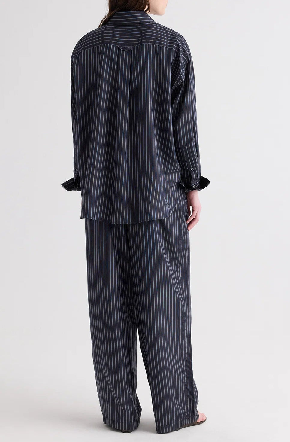 A person stands with their back to the camera, dressed in the TWP PJ Evening Stripe Silk Pant set—a loose-fitting, dark ensemble featuring wide-leg pants and a long-sleeved top adorned with a subtle evening stripe pattern.