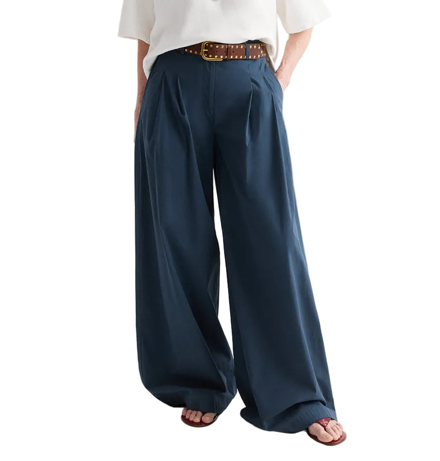 A person wearing the TWP Next Didi Raw Hem Pant—mid-rise, wide-leg navy blue cotton poplin pants—paired with a white top, a brown studded belt, and sandals, with hands in pockets.