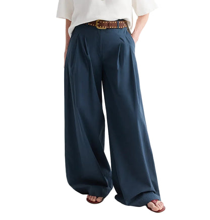 A person wearing the TWP Next Didi Raw Hem Pant—mid-rise, wide-leg navy blue cotton poplin pants—paired with a white top, a brown studded belt, and sandals, with hands in pockets.