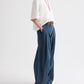 A person wearing a white short-sleeve cotton poplin top, the TWP Next Didi Raw Hem Pant in blue, a brown belt, a red necklace, and sandals, stands with hands in pockets against a plain background.