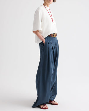 A person wearing a white short-sleeve cotton poplin top, the TWP Next Didi Raw Hem Pant in blue, a brown belt, a red necklace, and sandals, stands with hands in pockets against a plain background.
