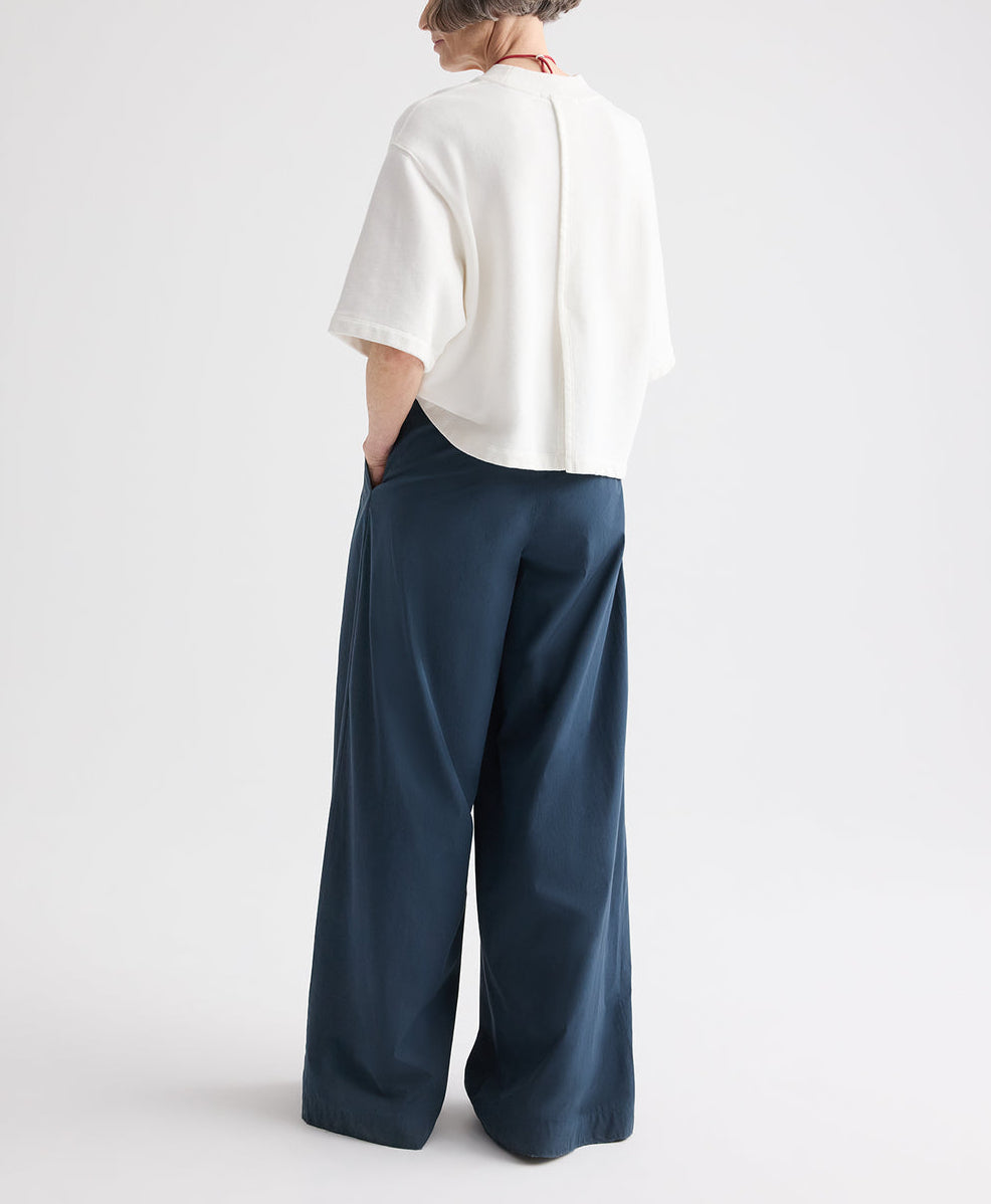 A person stands with hands in pockets, facing away from the camera against a plain white background, wearing a loose white top and the TWP Next Didi Raw Hem Pant — wide-leg, mid-rise navy blue cotton poplin pants.