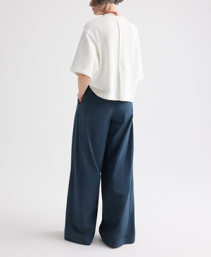 A person stands with hands in pockets, facing away from the camera against a plain white background, wearing a loose white top and the TWP Next Didi Raw Hem Pant — wide-leg, mid-rise navy blue cotton poplin pants.