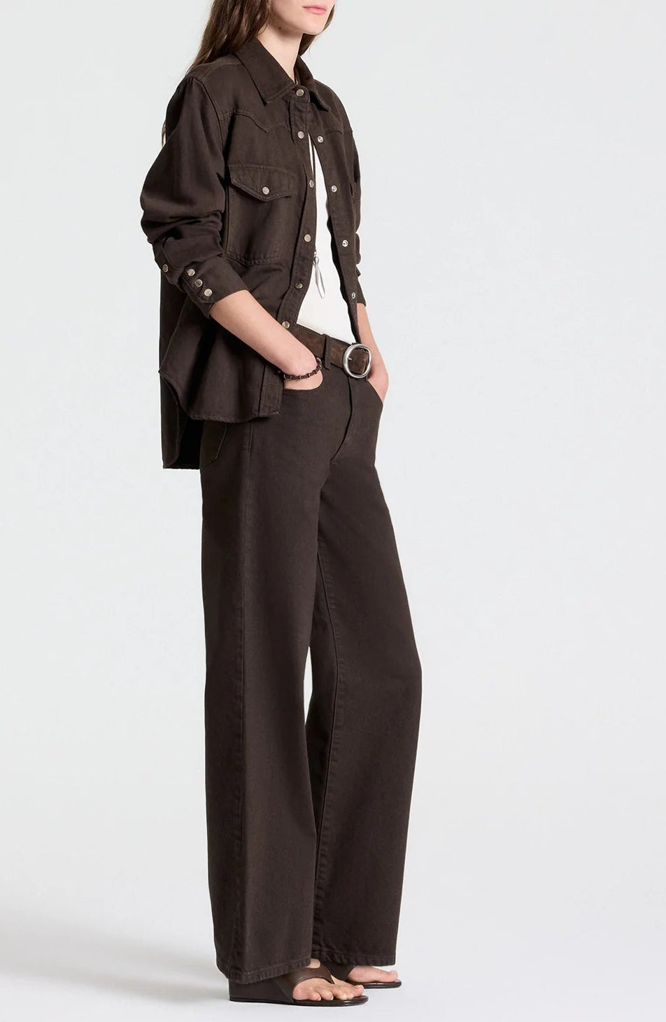 A person wearing a dark brown button-down shirt and matching dark chocolate TWP Harvest Moon Jean wide-leg jeans with a black belt and sandals.