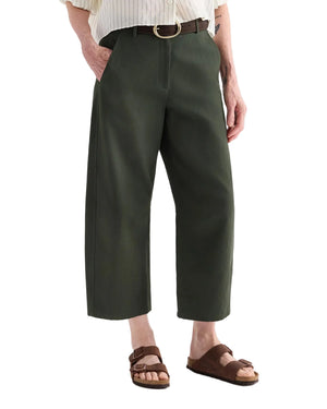 A person wears TWP Slouchy BF Cotton Linen Twill Trousers in dark green with a light shirt, black belt, and brown sandals, standing hands in pockets.