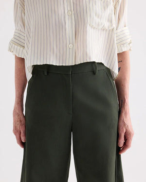 A person wears TWP's Slouchy BF Cotton Linen Twill Trouser in dark green, paired with a cream-striped button-up shirt, shown from shoulders to knees with arms relaxed at their sides.