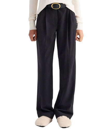 A person wearing the TWP Ivy Pant, black mid-rise pleated trousers crafted from luxurious Italian wool twill, styled with a black belt, white long-sleeve top, and white slippers, standing against a white background.