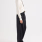 A person stands sideways wearing a white long-sleeve shirt, TWP’s Ivy Pant—mid-rise pleated Italian wool twill trousers in Smoke—and slippers, accessorized with a necklace and a belt featuring a large buckle.