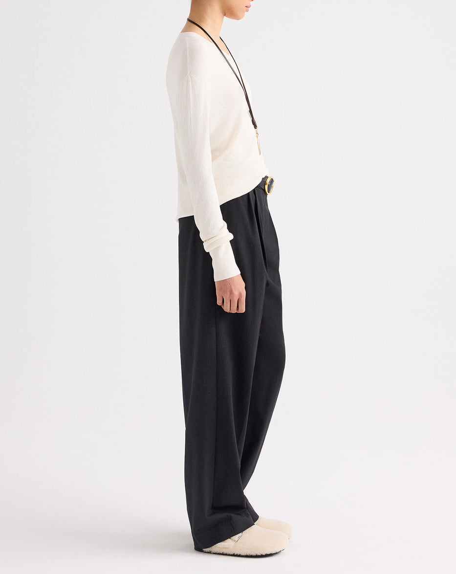 A person stands sideways wearing a white long-sleeve shirt, TWP’s Ivy Pant—mid-rise pleated Italian wool twill trousers in Smoke—and slippers, accessorized with a necklace and a belt featuring a large buckle.