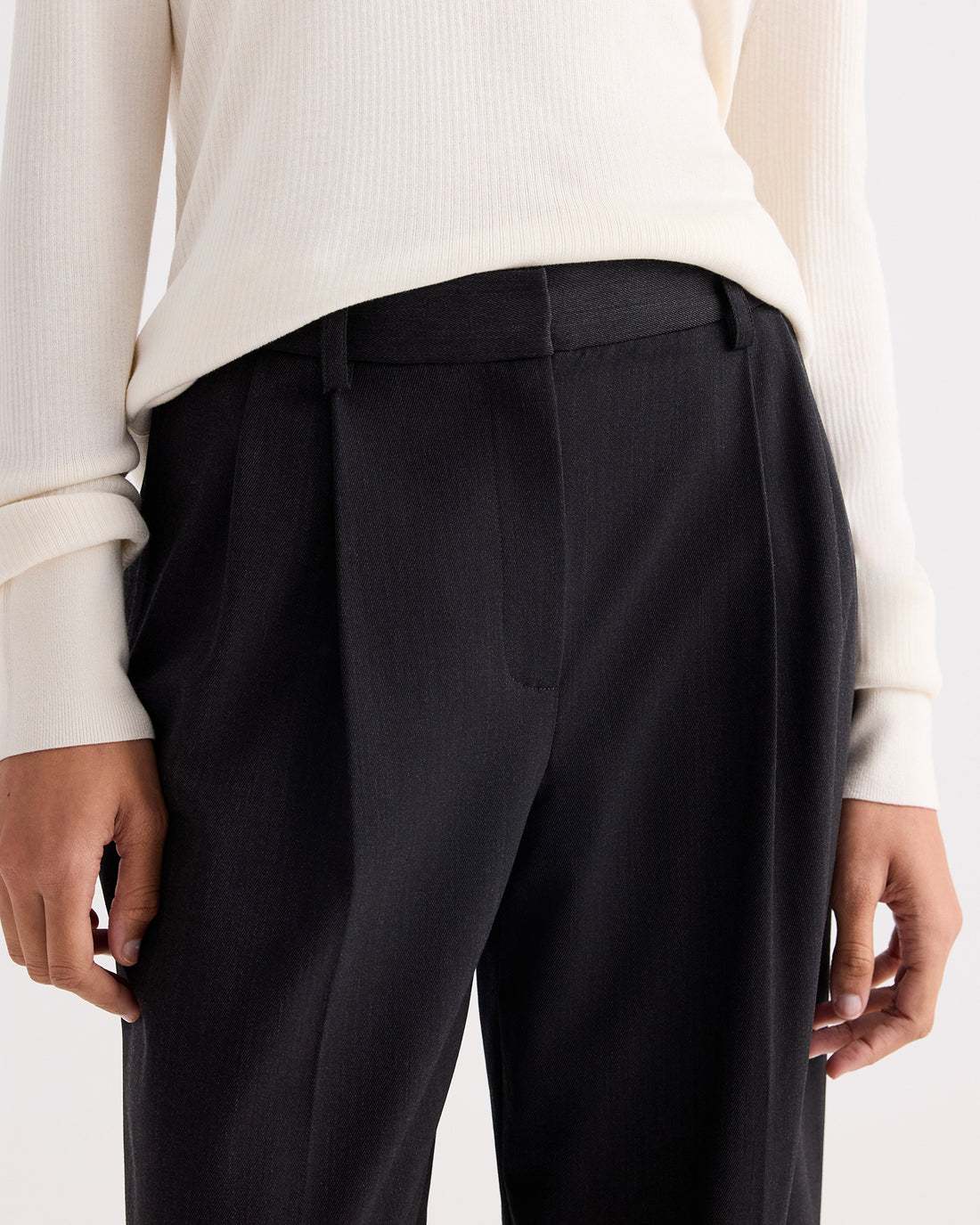 A person wearing a cream long-sleeve ribbed top tucked into the TWP Ivy Pant—high-waisted black Italian wool twill pleated trousers—shown from mid-torso to upper thighs.