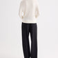 A person stands with their back to the camera, wearing a long-sleeve white top and the Ivy Pant by TWP—loose mid-rise pleated trousers crafted from Italian wool twill—set against a plain white background.