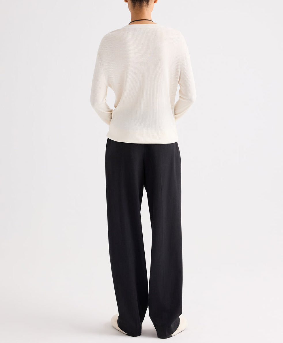 A person stands with their back to the camera, wearing a long-sleeve white top and the Ivy Pant by TWP—loose mid-rise pleated trousers crafted from Italian wool twill—set against a plain white background.