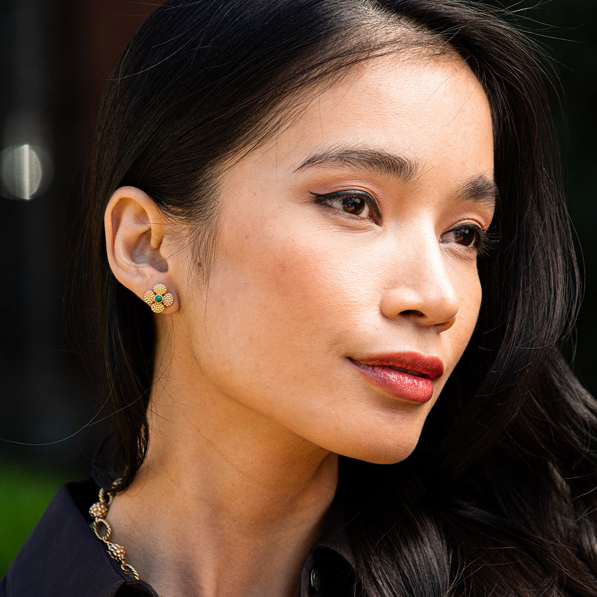 A woman with long dark hair wears Juliska Capucine De Wulf Berry Clover Convertible Duo Earrings and a black top, looking to the side outdoors.