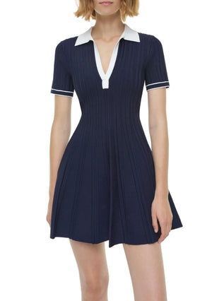 A woman models the Simkhai Page Knit Polo Mini Dress, featuring short sleeves, navy blue knit fabric, a white collar, and white sleeve trim, set against a plain background.