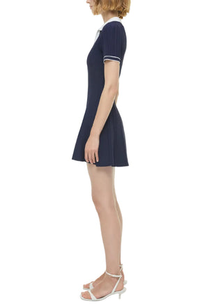 A woman stands in profile wearing the Simkhai Page Knit Polo Mini Dress in navy with a white collar and trim, short sleeves, and white heeled sandals.