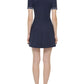 A woman stands facing away, wearing the Simkhai Page Knit Polo Mini Dress in navy blue with a flared skirt and white-heeled sandals.