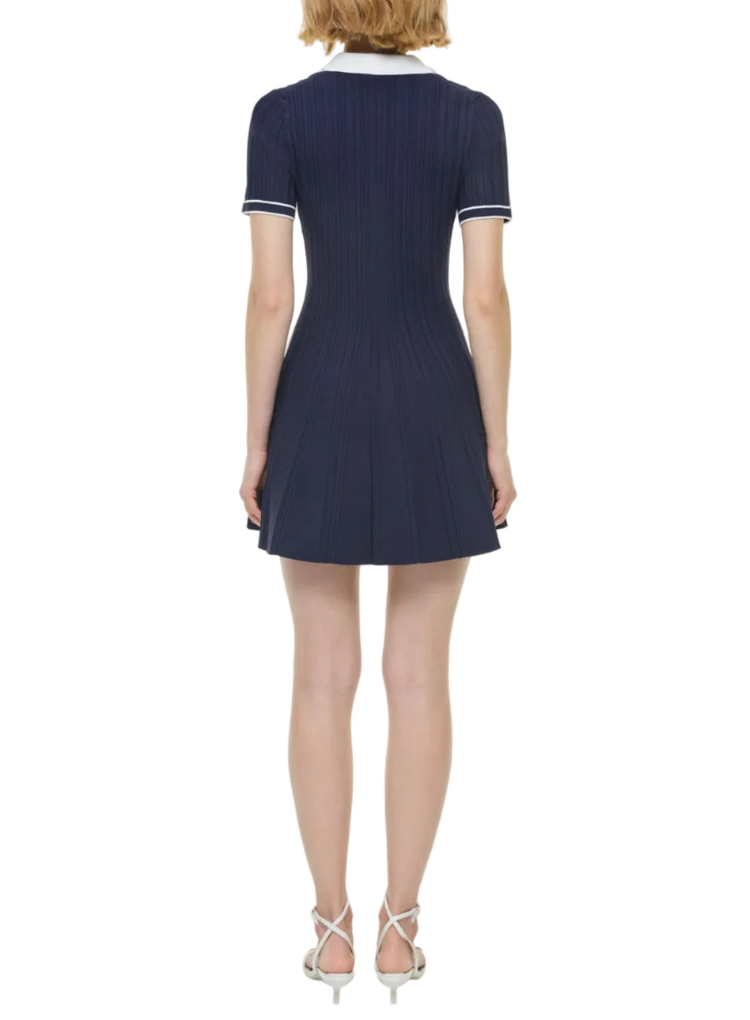 A woman stands facing away, wearing the Simkhai Page Knit Polo Mini Dress in navy blue with a flared skirt and white-heeled sandals.