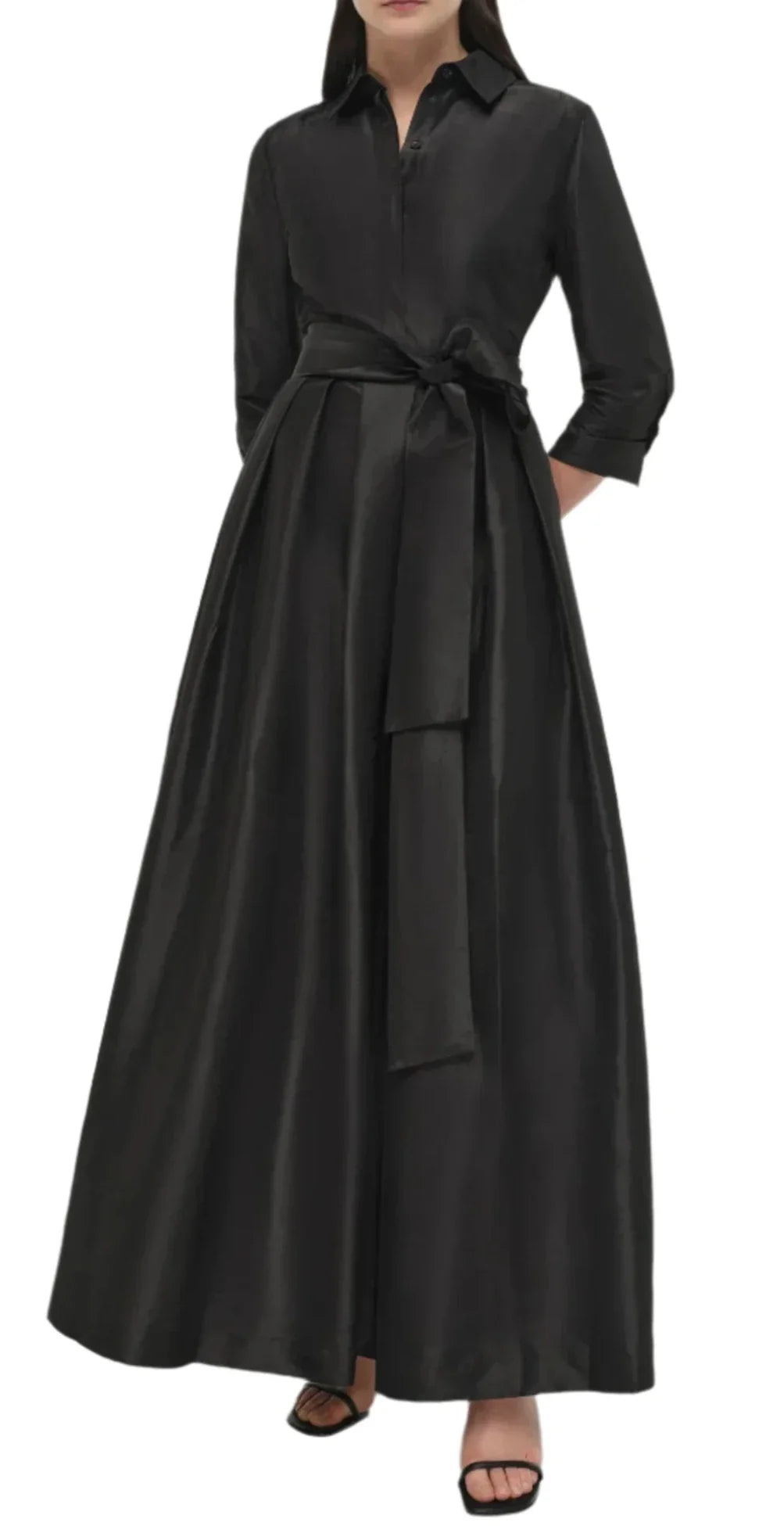 Woman wearing a long, black Simkhai Julip Belted Gown with tailored details, featuring a tied waist belt and long sleeves. She is standing with her hands at her sides and is wearing black open-toe sandals.