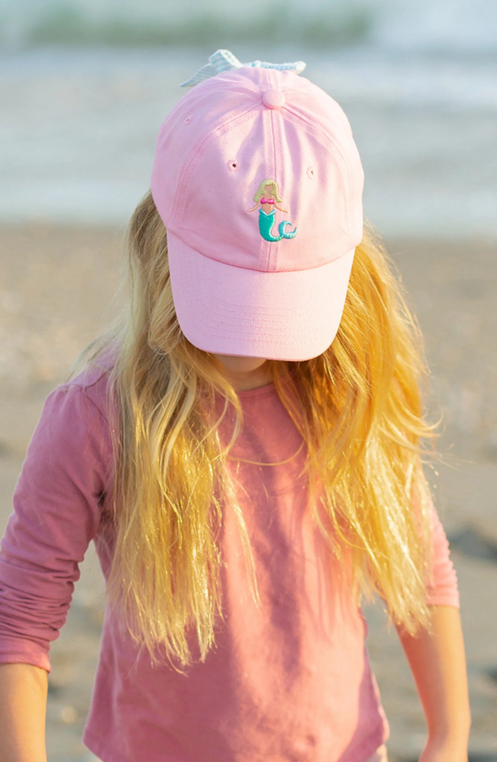 A child with long blonde hair wears a pink shirt and the Blonde Mermaid Bow Baseball Hat by Bits & Bows, standing outdoors and looking down—perfect for summer sportswear.