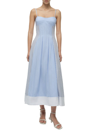 A woman wears the Simkhai Chamberlain Cotton Midi Dress in periwinkle multi stripe with a cami neckline and white heeled sandals, standing against a plain white background.