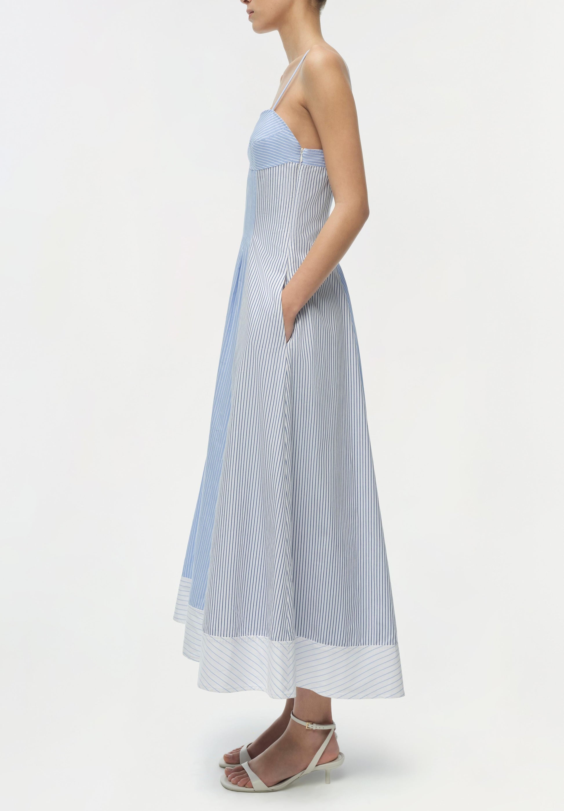 A woman in the Simkhai Chamberlain Cotton Midi Dress with multi stripes and a cami neckline stands sideways, hands in pockets, paired with light pink heeled sandals.