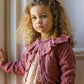 A young girl with curly blonde hair stands indoors by a window, wearing the Noralee Baby Girls' and Girls' Ruffle Cardigan over a floral dress, looking at the camera with a neutral expression.