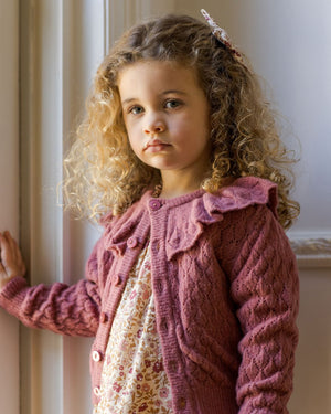 A young girl with curly blonde hair stands indoors by a window, wearing the Noralee Baby Girls' and Girls' Ruffle Cardigan over a floral dress, looking at the camera with a neutral expression.