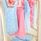 Children’s clothes in blue and pink patterns, including The Beaufort Bonnet Company’s Baby Stillman Sunsuit, hang on wooden hangers on a bamboo rack, with matching folded garments neatly arranged below.