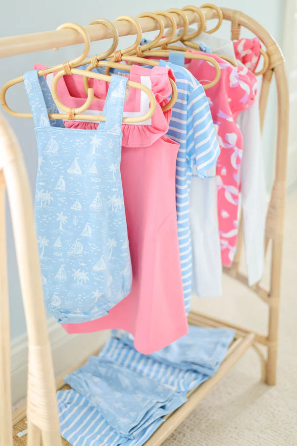 Children’s clothes in blue and pink patterns, including The Beaufort Bonnet Company’s Baby Stillman Sunsuit, hang on wooden hangers on a bamboo rack, with matching folded garments neatly arranged below.
